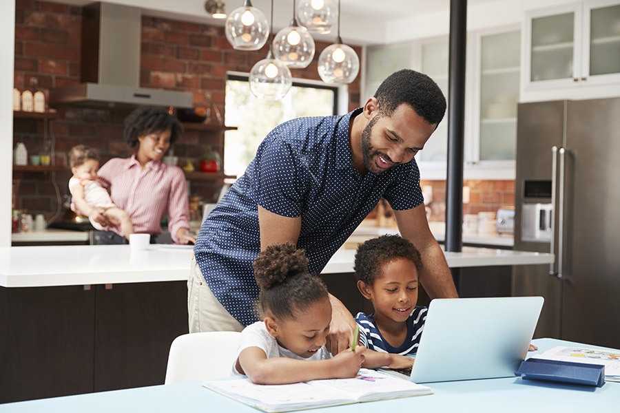 Personal Insurance - A Father is Helping His Two Young Children With Homework While the Mother is Holding a Baby and Checking Her Laptop in the Kitchen